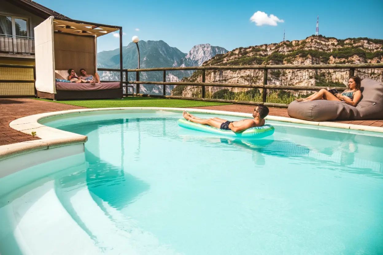 La piscina panoramica con gli ospiti sul materassino e sui pouf, dietro le montagne dell'Alto Garda